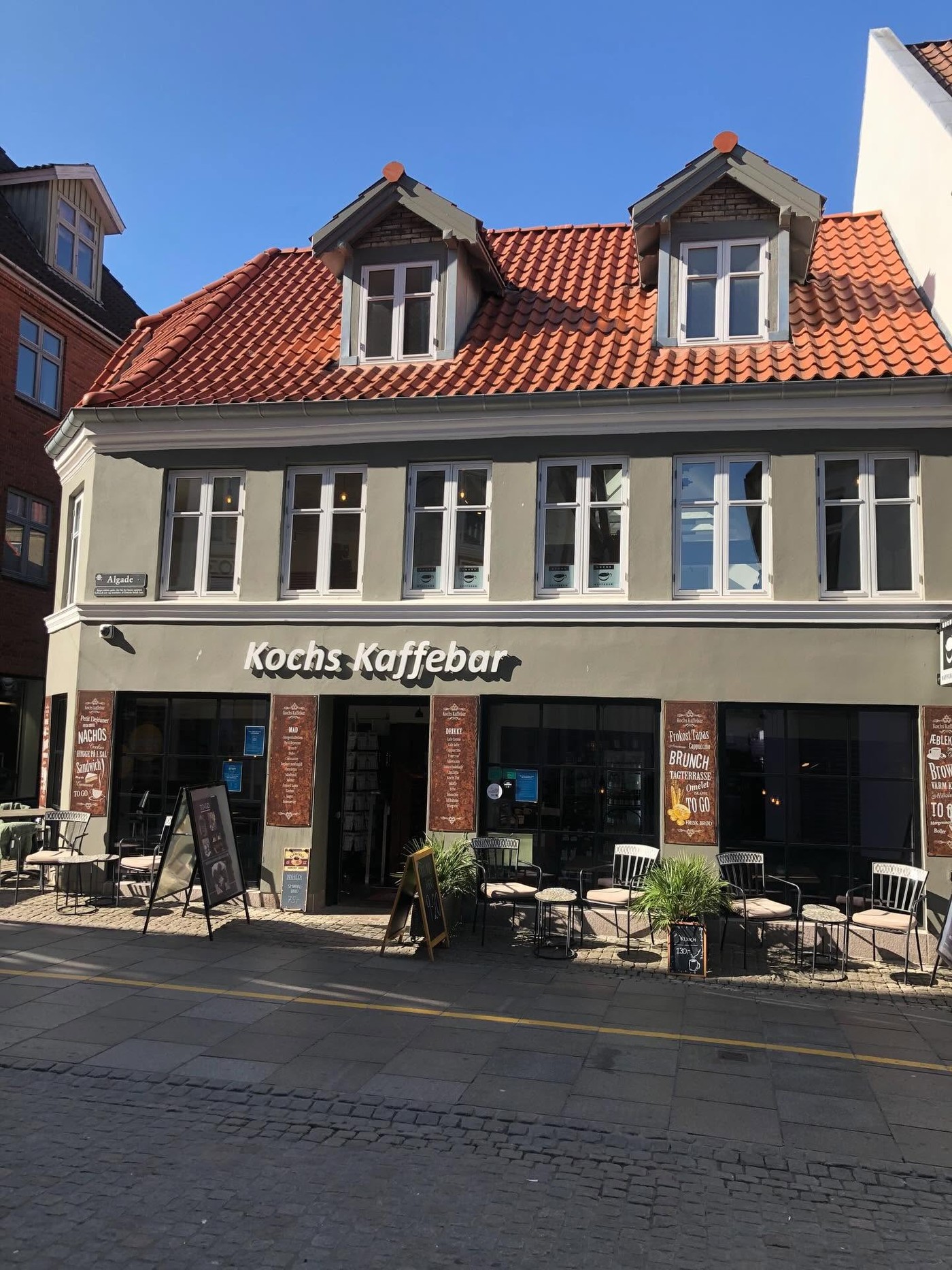 The gray facade of Kochs Kaffebar with red tiled dormers above and outdoor cafe seating and menu boards along the sidewalk