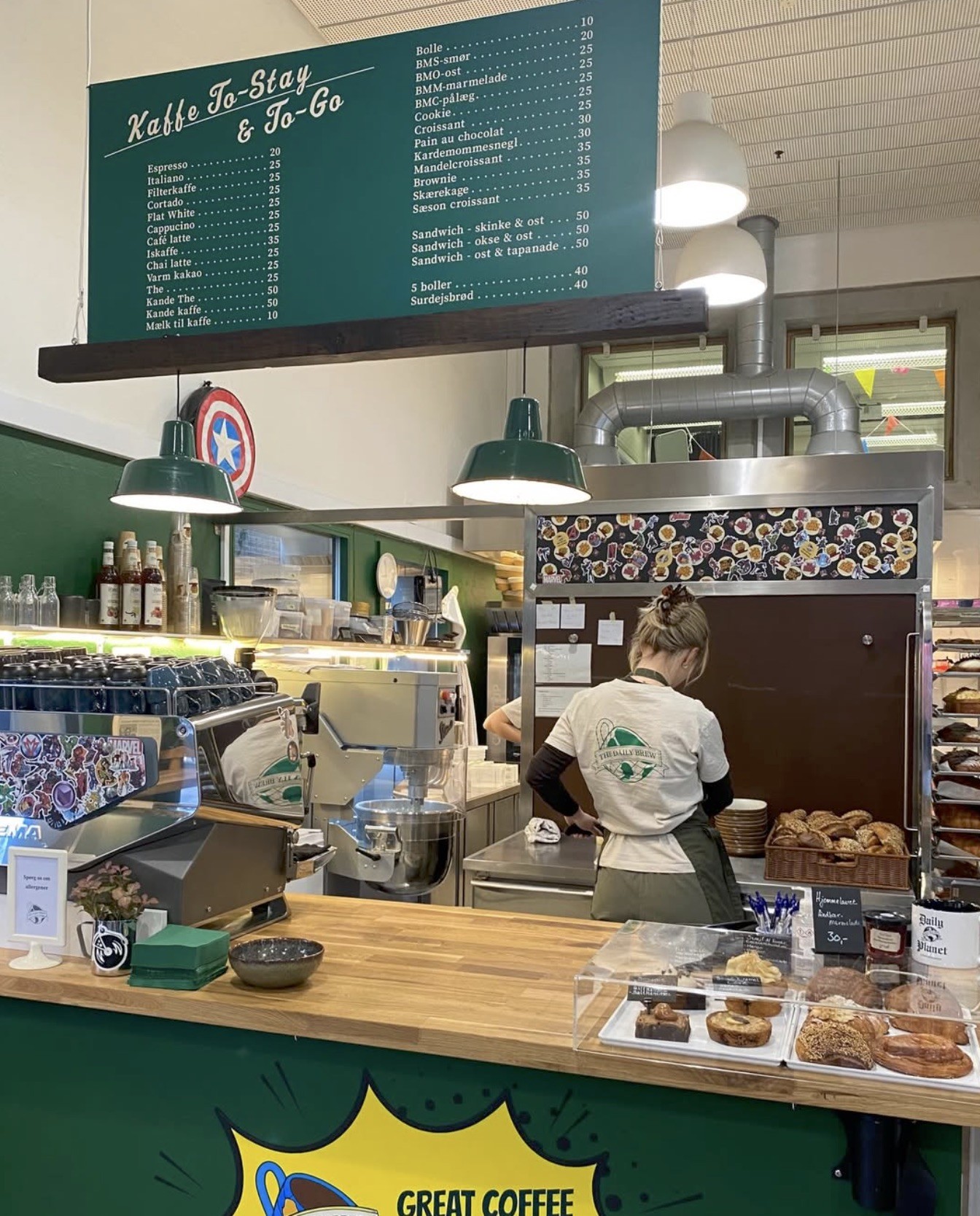 A barista in a The Daily Brew shirt working behind the counter, with a chalkboard coffee menu overhead and pastries in a case up front