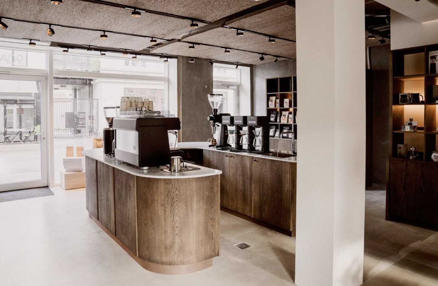 A curved wood counter with an espresso machine and grinders beneath an acoustic panel ceiling at Europa Kaffe and Te
