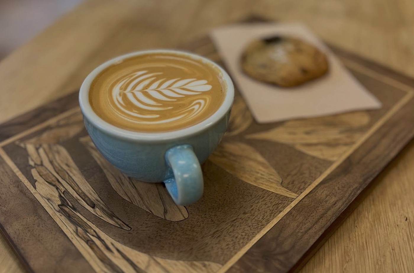 A cappuccino with rosetta latte art beside a chocolate chip cookie on an inlaid wood tray at Stiller's Coffee