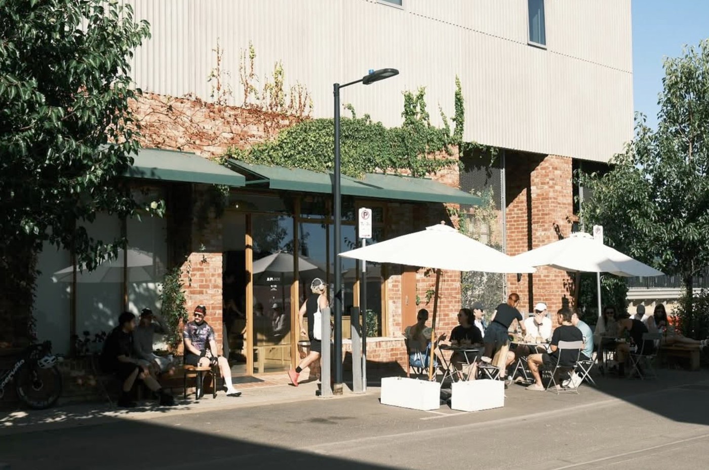 The brick facade of A Place Coffee with green awnings, ivy climbing the walls, and cyclists gathered at outdoor tables under umbrellas