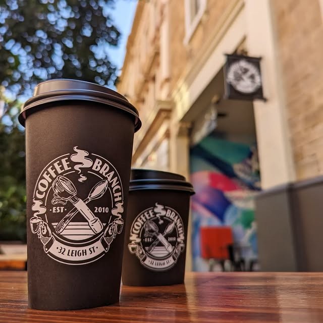 Two branded Coffee Branch takeaway cups on an outdoor table, with the Leigh Street storefront and mural in the background.