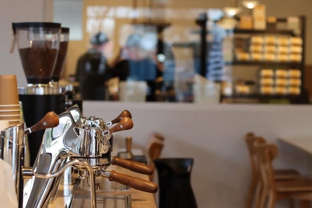 Espresso machine group heads in the foreground at Elementary Coffee, with the pastry case and counter staff blurred in the background.