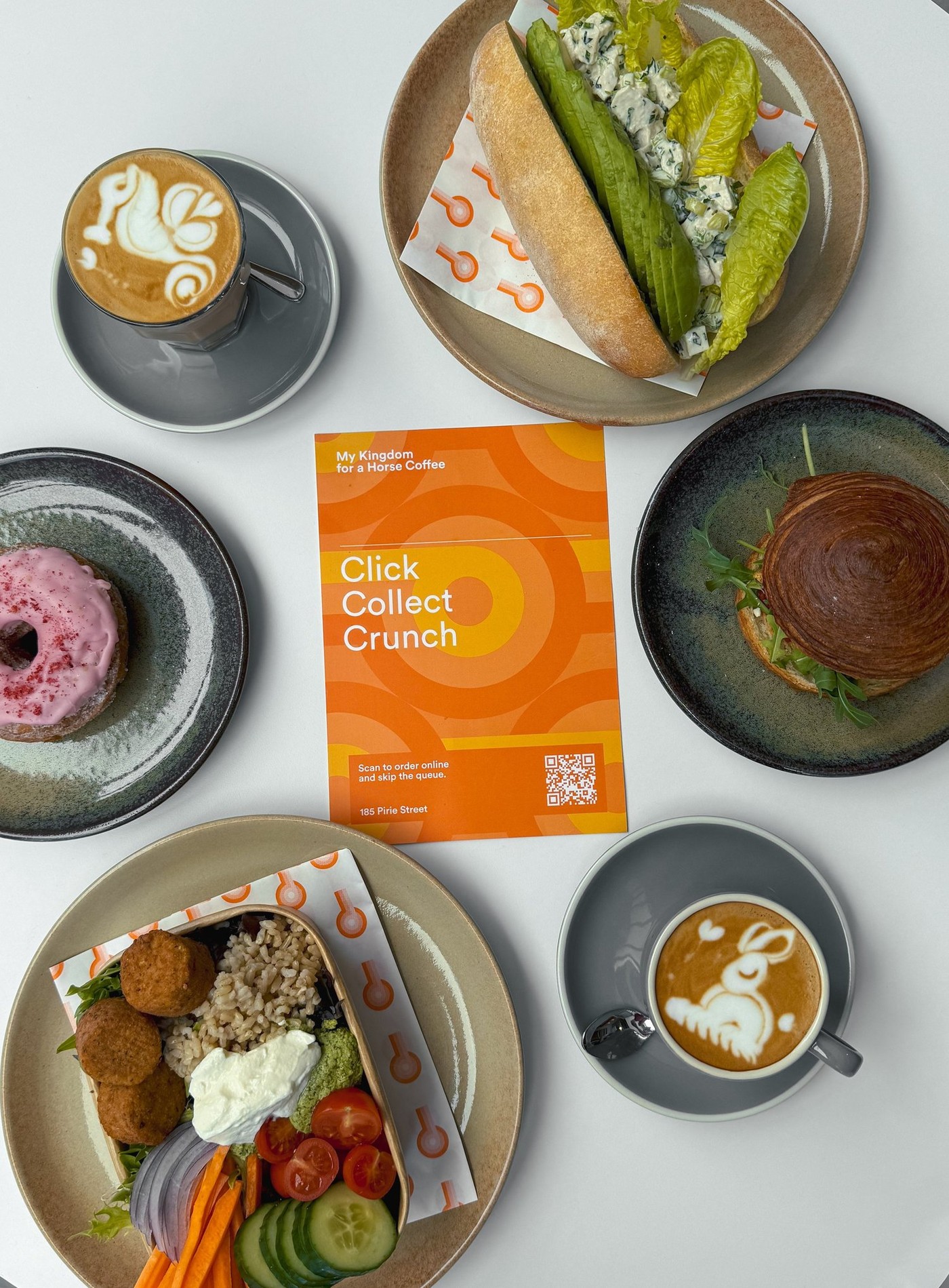 A flat lay of latte art, a croissant sandwich, a falafel bowl and a sandwich around a branded My Kingdom for a Horse Coffee order card listing the 185 Pirie Street address.