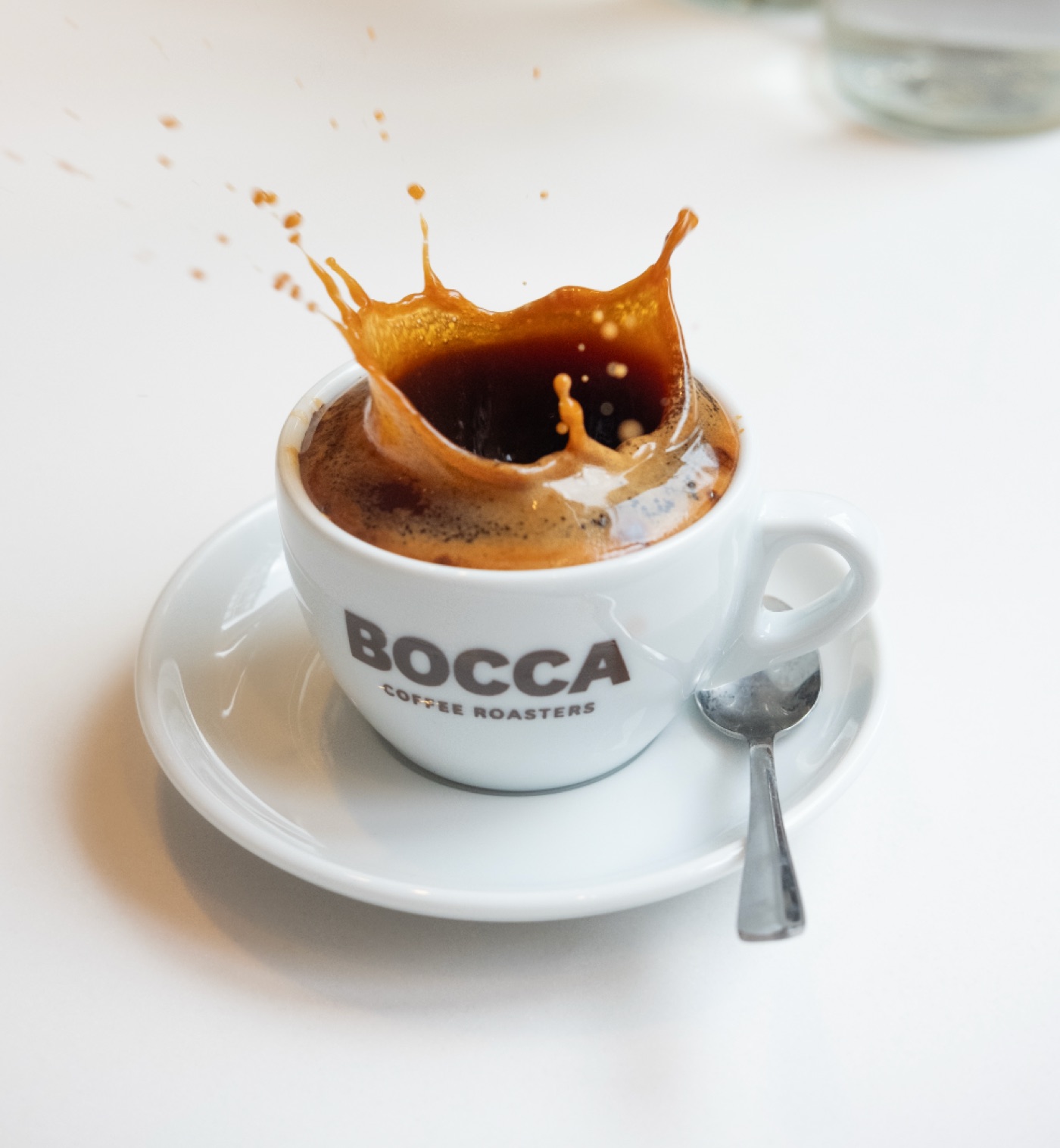 An espresso splashing out of a white cup branded Bocca Coffee Roasters, on a matching saucer with a spoon