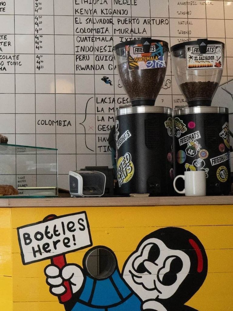 Twin grinders loaded with branded beans beside a handwritten chalkboard menu, a Bottles Here mascot mural below