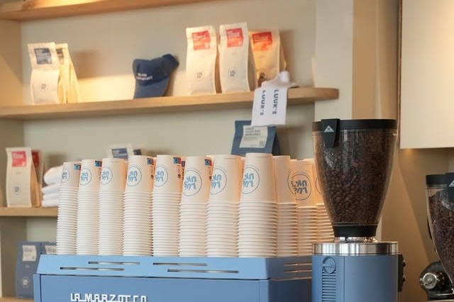 The blue La Marzocco espresso machine at Luuk's Coffee Noordermarkt with branded cups stacked on top and retail coffee bags on the shelf behind