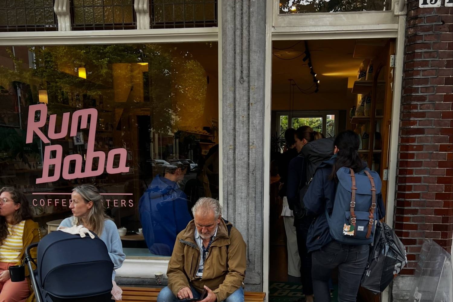 The Rum Baba Coffee Roasters storefront window with the shop's pink lettering, customers seated on the bench out front