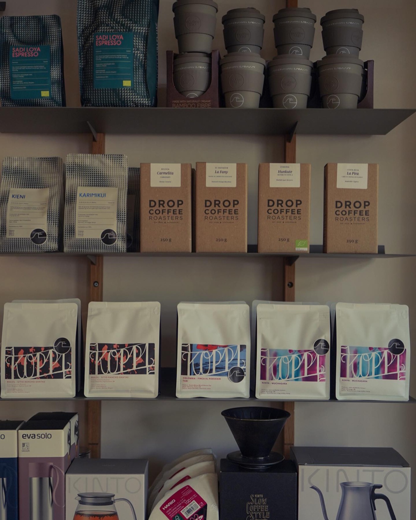 A retail shelf of bagged beans from Drop Coffee and Koppi, reusable cups, and brewing gear at Scandinavian Embassy