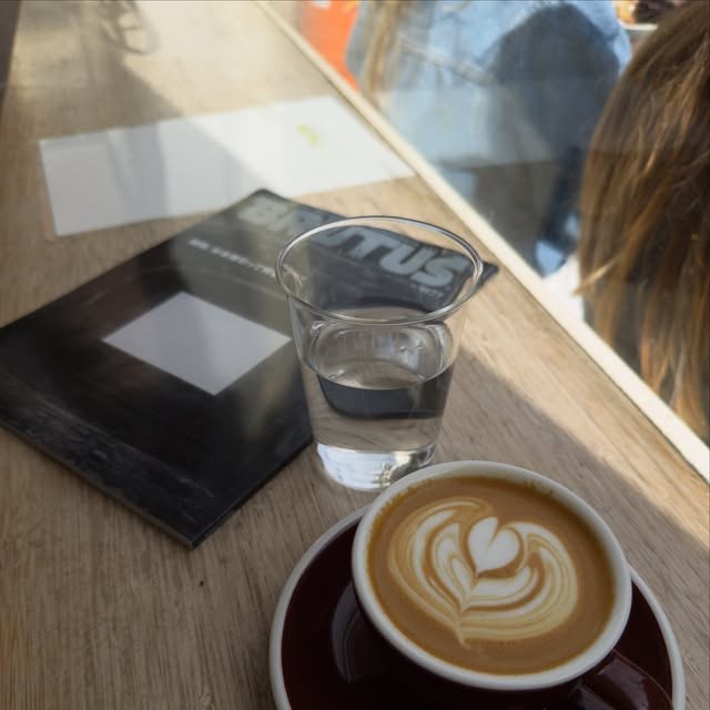 A cappuccino with latte art beside a glass of water and a magazine on the sunlit wooden counter