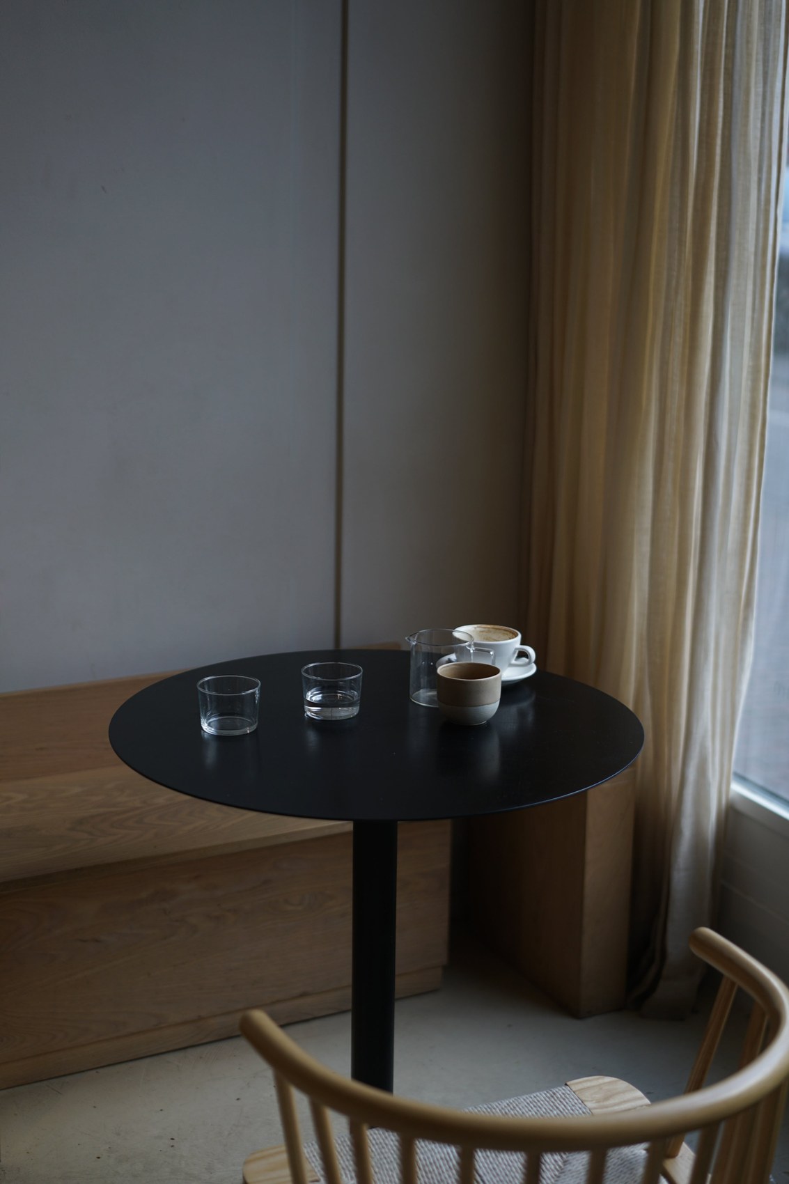 A round black table set with water glasses, a ceramic cup and a cappuccino beside linen curtains