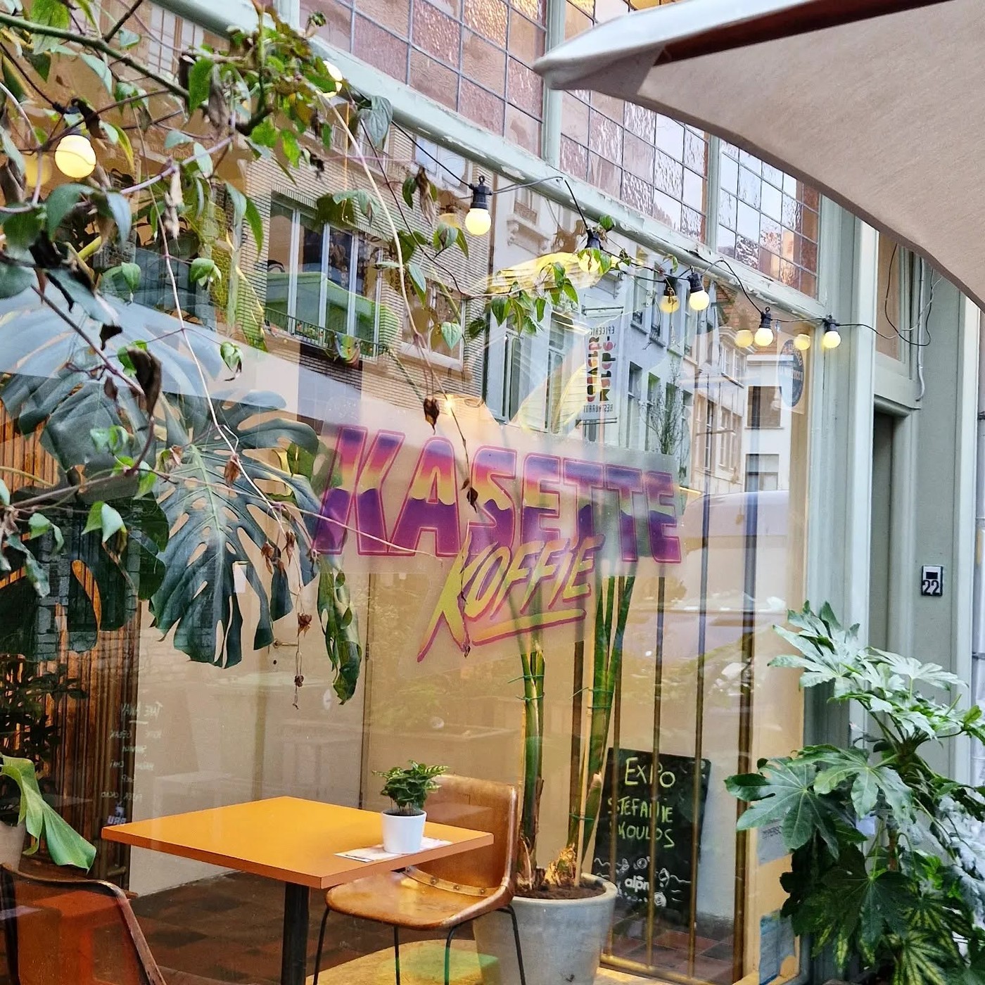 The Kasette Koffie retro lettering painted on the front window above a small orange table and potted plants