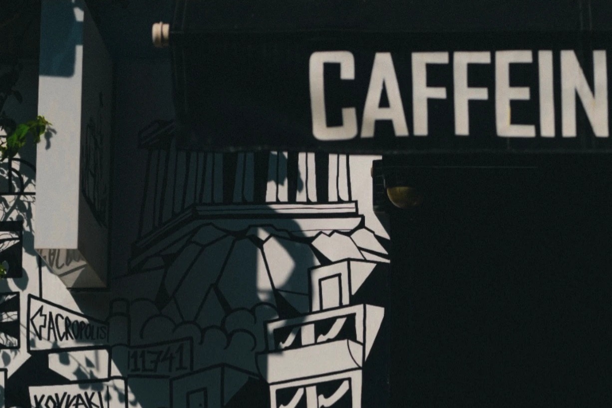 The black and white Koukaki skyline mural painted above the CAFFEINE sign on Caffeine Dealer's storefront