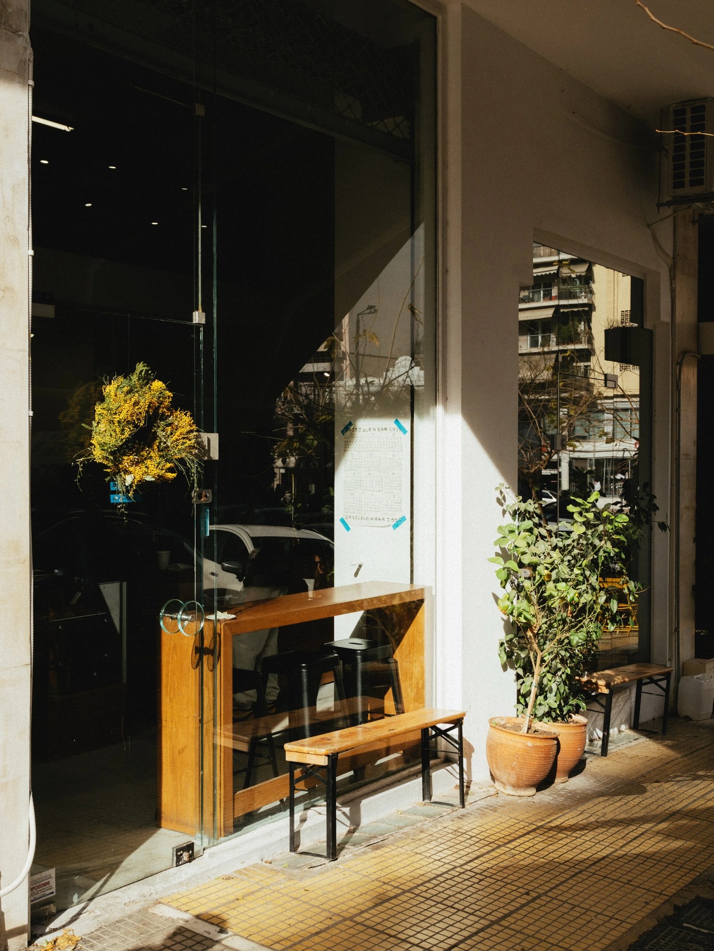 The sunlit glass storefront of Foyer Espresso Bar with a wooden bench and potted plants out front