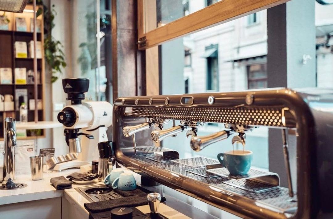 A three group espresso machine pulling a shot beside shelves of retail coffee bags at Samba Coffee Roasters