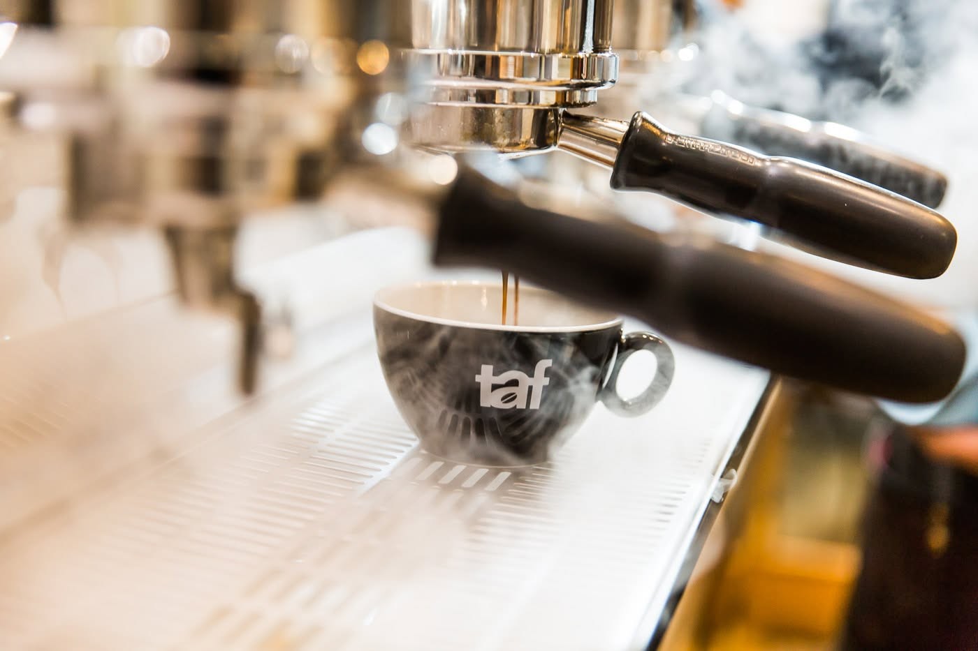 Espresso pouring into a branded TAF cup under the group head of an espresso machine, steam rising