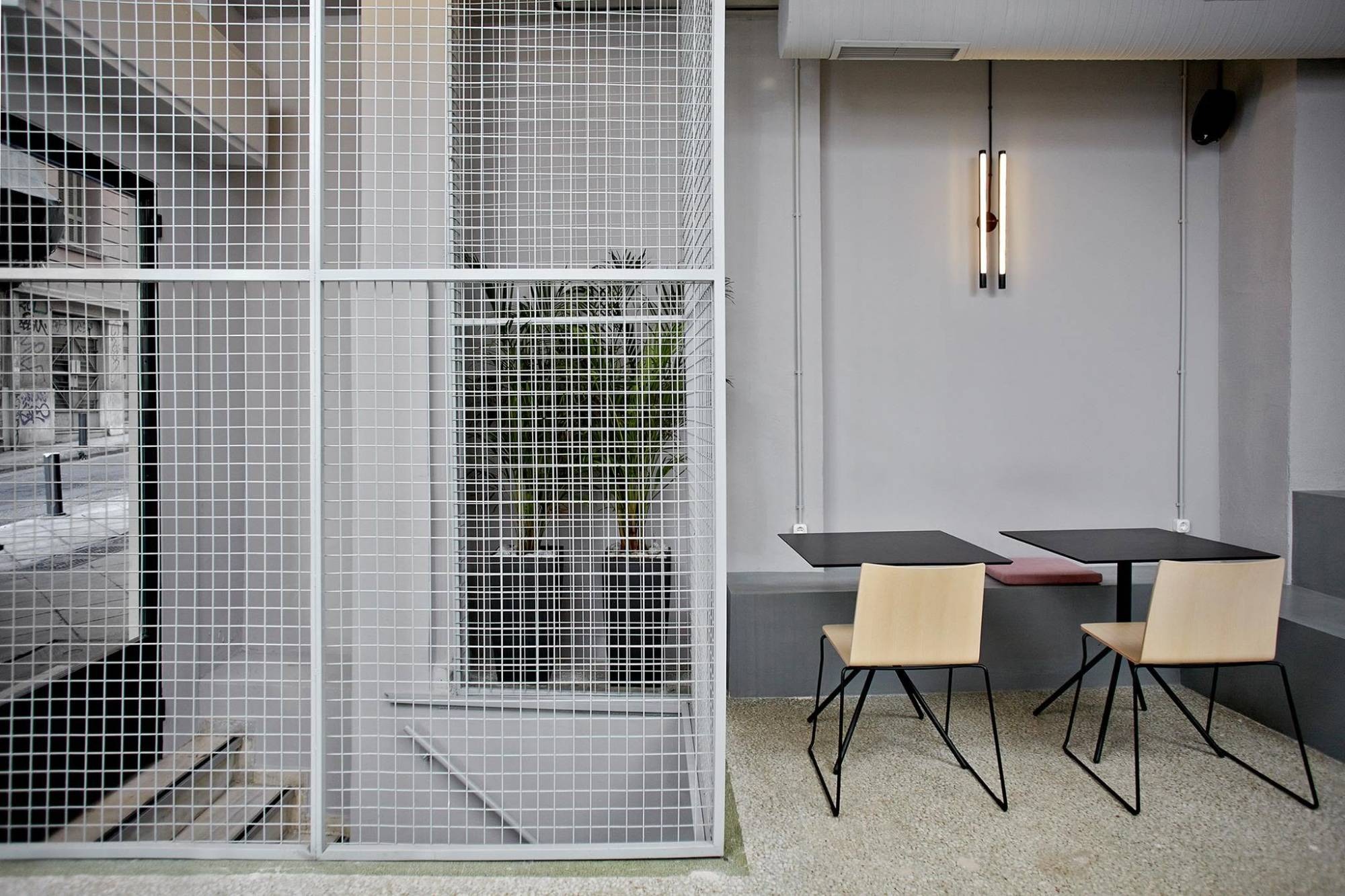 THIRD PLACE.'s minimalist seating nook, wire mesh screening a plant filled stairwell beside two small tables and pale wood chairs