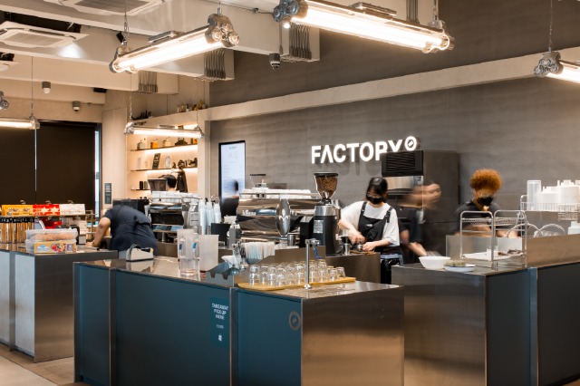 Baristas at work behind the counter at Factory Coffee's Phaya Thai branch, under a glowing FACTORY wall sign