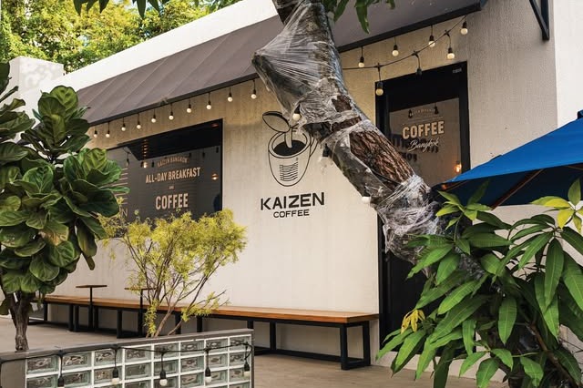 The white exterior of Kaizen Coffee Ekkamai with a large KAIZEN COFFEE wall sign, string lights, wooden benches, and potted plants out front