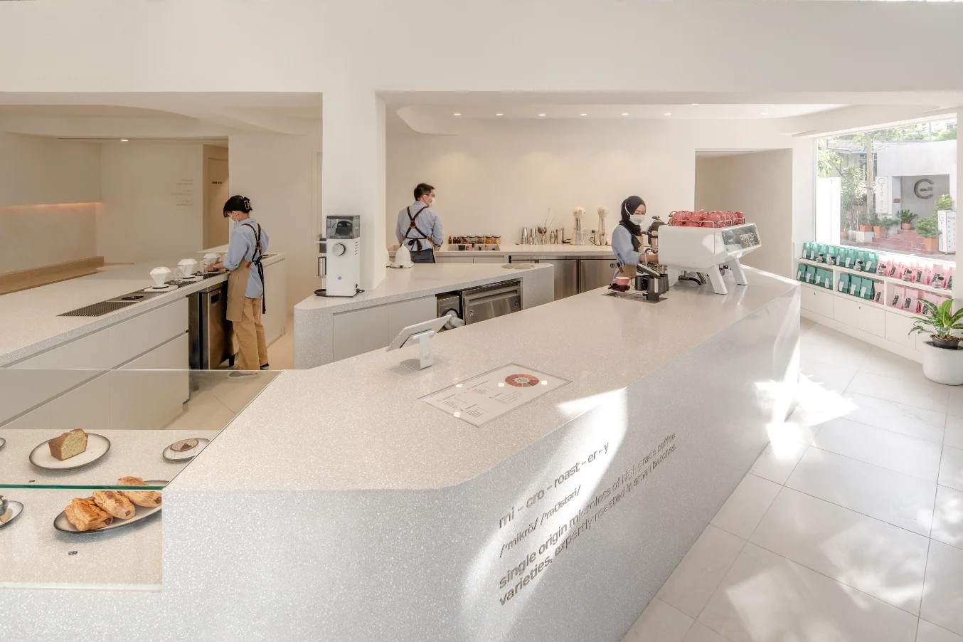 The white terrazzo counter at PAGA Microroastery with micro-roastery lettering along its side, baristas at work, a shelf of bagged beans by the window and a glass-fronted roasting room above