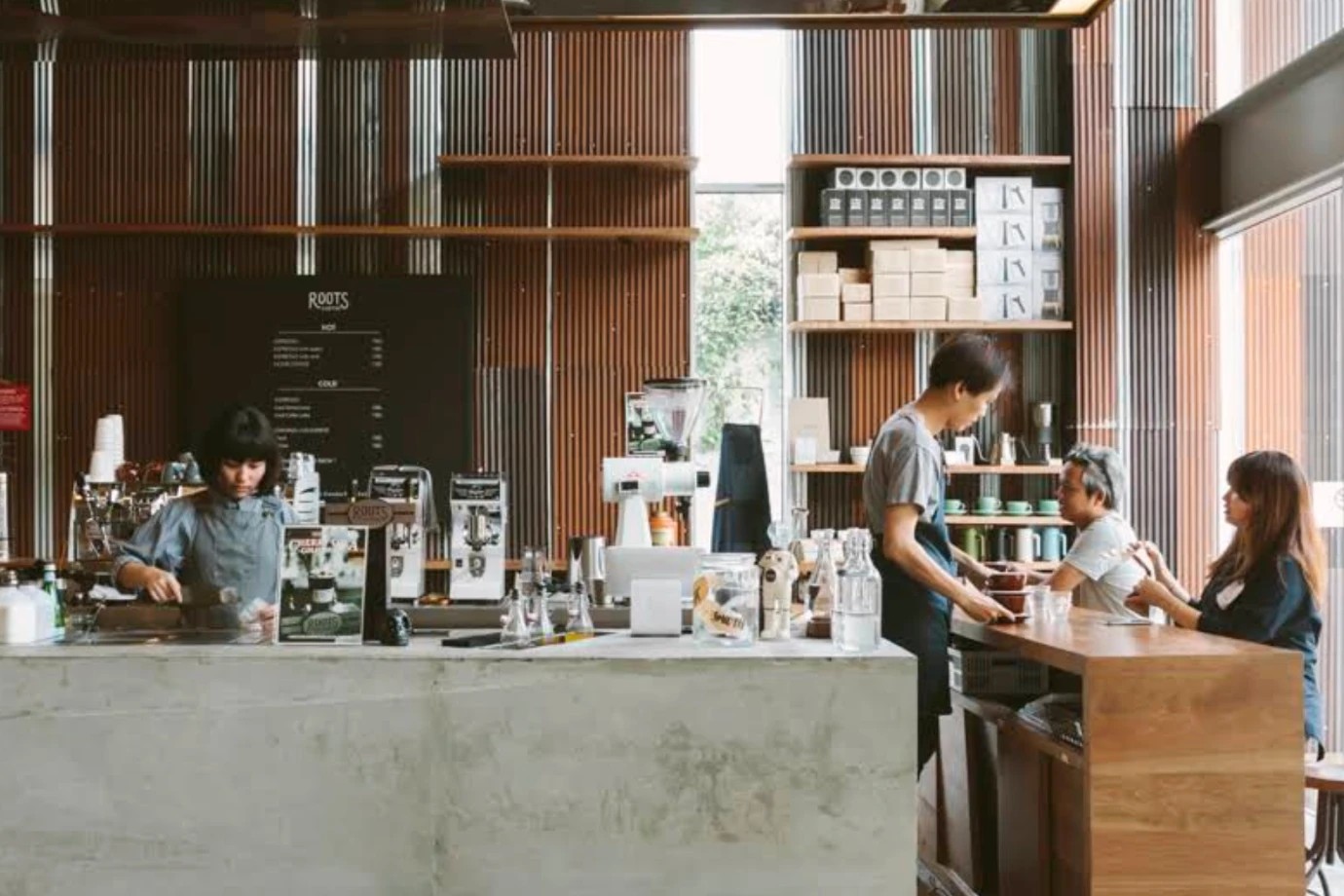 The open air counter at Roots at the Commons, a concrete bar with a ROOTS menu board behind it, a barista serving guests at a wood bar and shelves of brewing gear against the corrugated metal wall
