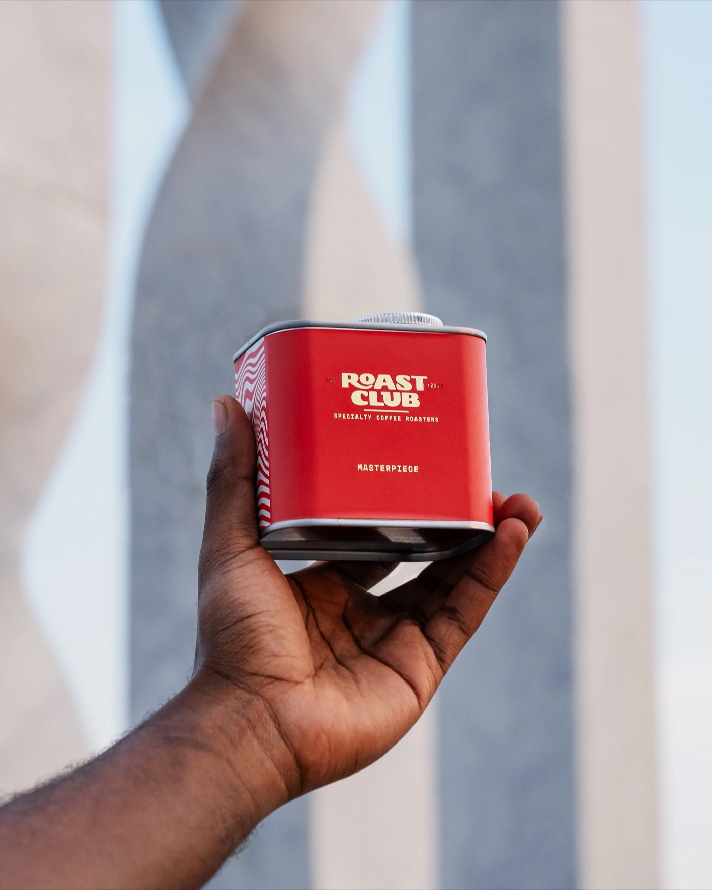 A hand holding a red Roast Club Masterpiece coffee tin against a sunlit stone backdrop
