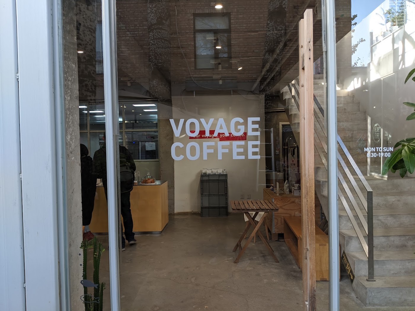The VOYAGE COFFEE wall lettering seen through the entrance, a silhouetted figure inside and a staircase leading up outside