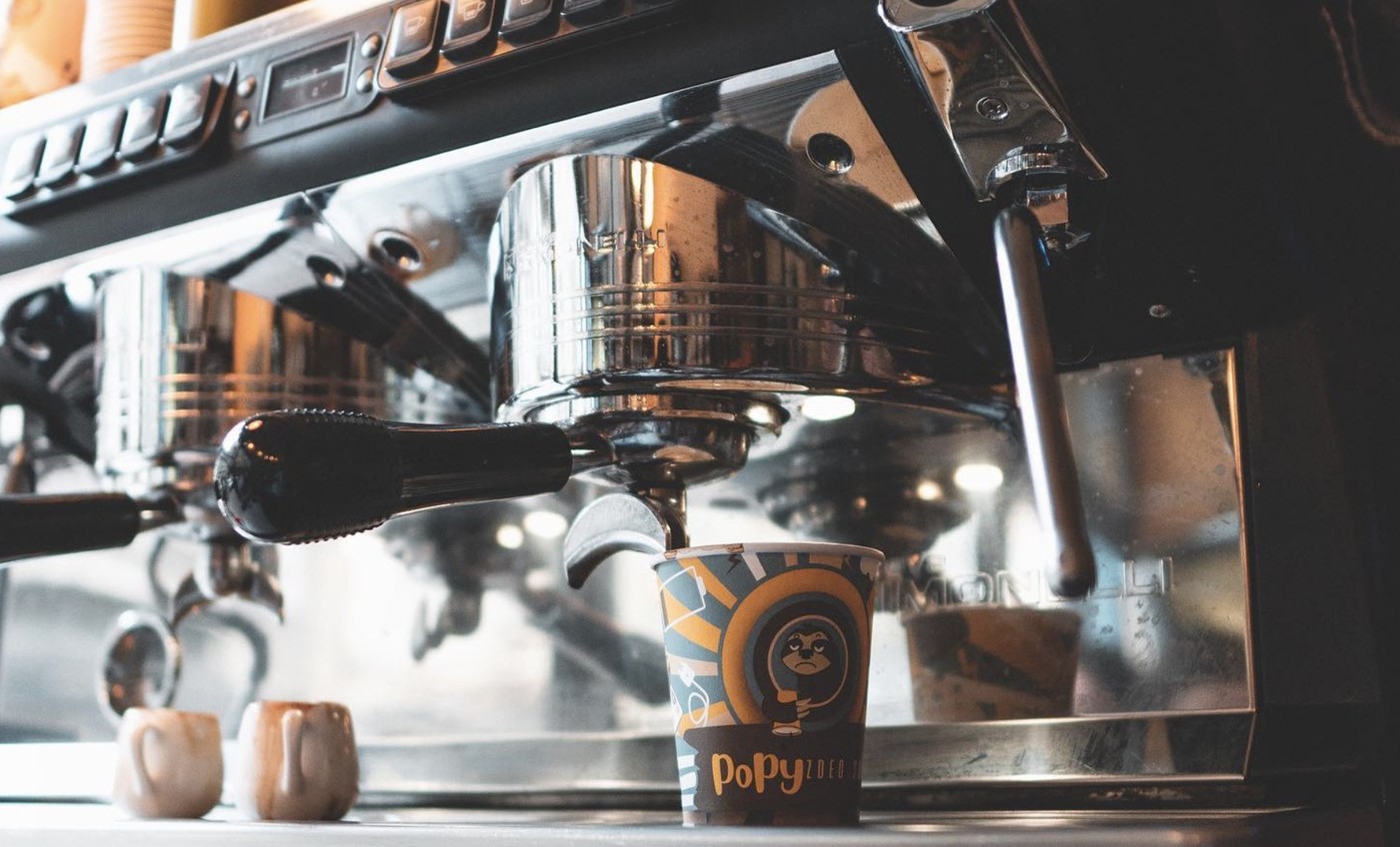 A branded Popy paper cup under the group head of an espresso machine, pulling a shot