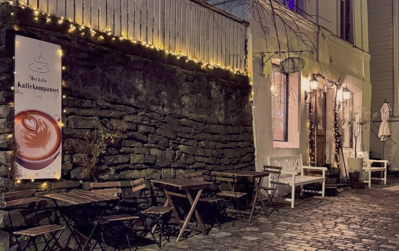 String lights and a latte art sign lining the stone alley outside Det Lille Kaffekompaniet at night