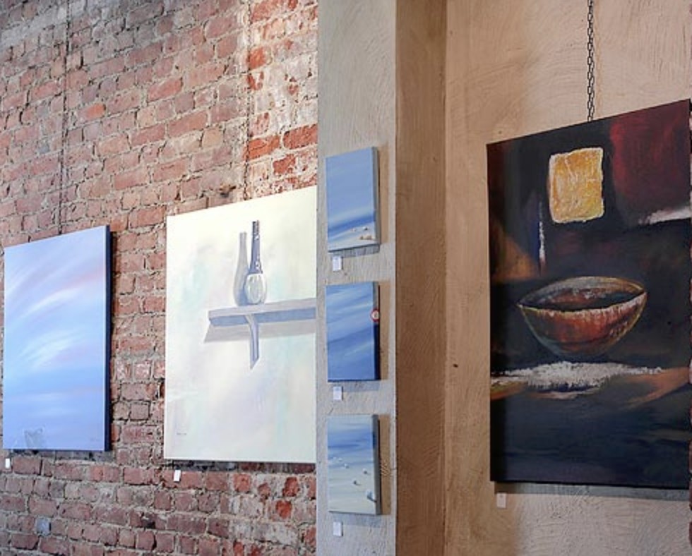 Paintings of varying sizes hung on exposed brick and wood walls in the Pygmalion gallery room