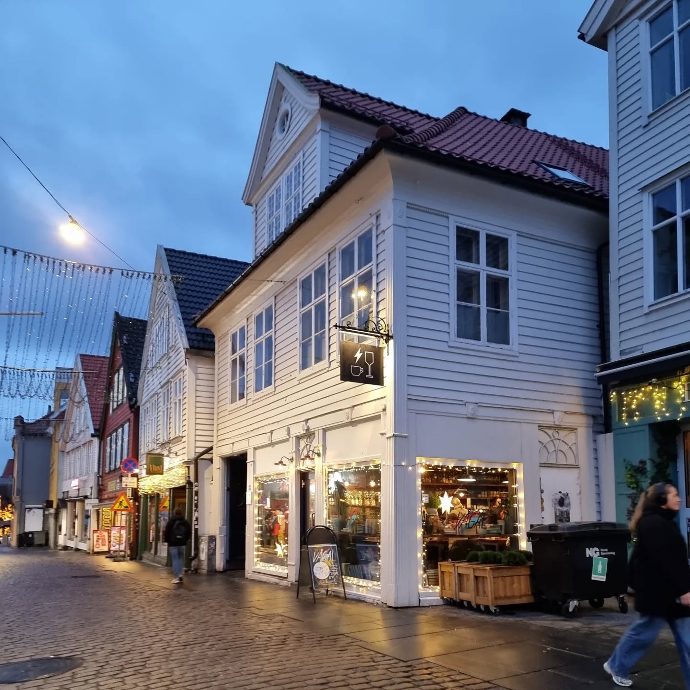 The white timber facade of Vågal at dusk, its coffee-and-wine glass sign lit above the illuminated storefront window