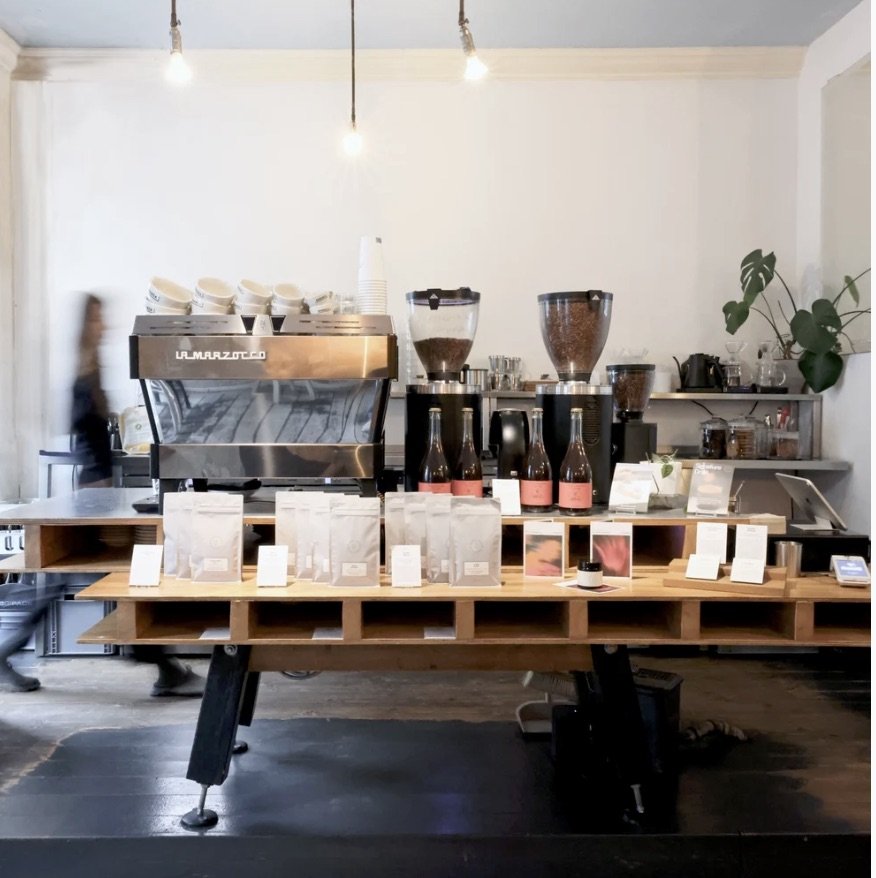 The retail counter at Bonanza Coffee, La Marzocco espresso machine, bagged beans on a wooden shelf, and a plant in the corner