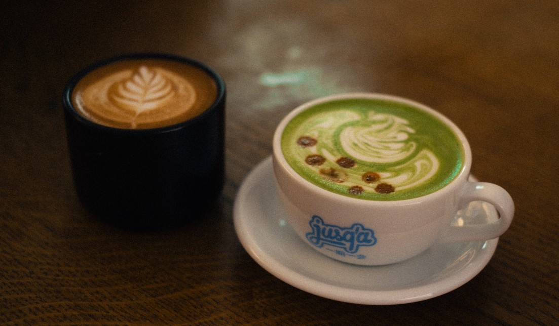 A cappuccino with leaf latte art beside a matcha latte in a branded jusq'à cup