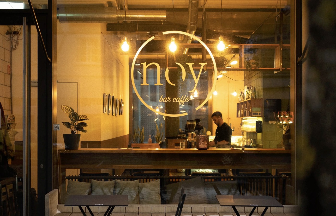 The illuminated noy bar caffè logo on the window, with the wooden counter and seating visible inside at night