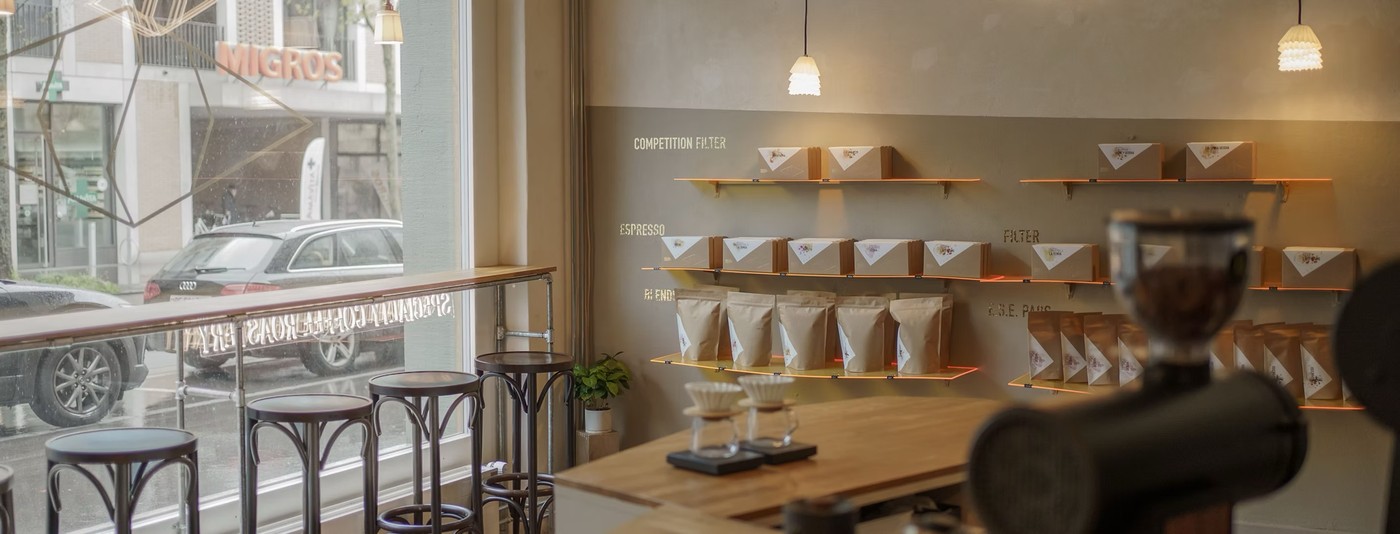 Retail bags of coffee organized by brew method on illuminated shelves along the wall at Ojo de Café's roastery showroom