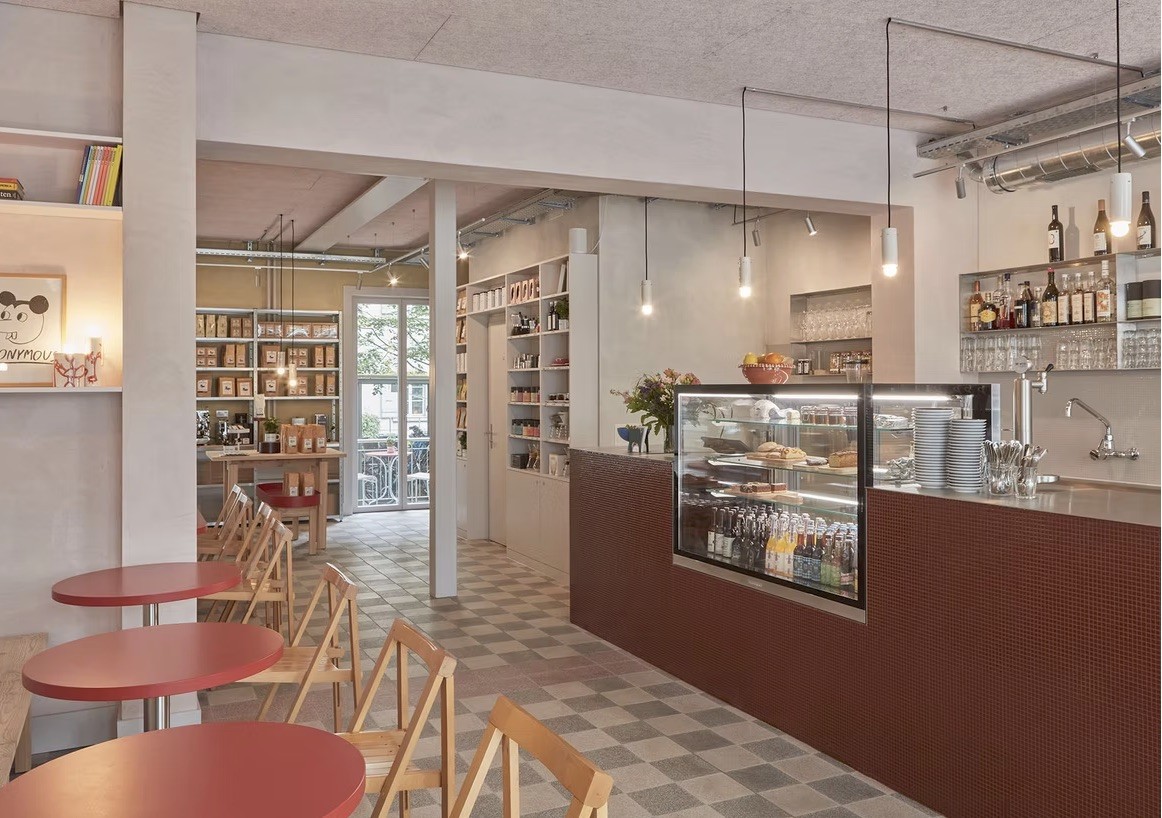 The tiled counter and pastry case at RÖSTEREI, with retail coffee bags and brewing gear on shelves behind it and red cafe tables in the foreground