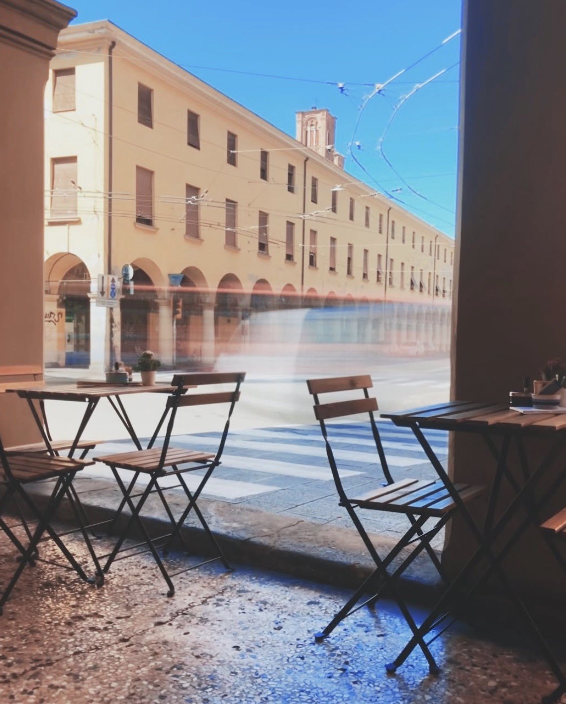 Empty bistro tables on Lampadina's covered terrace looking out at the porticoed street with a passing tram blurred in motion