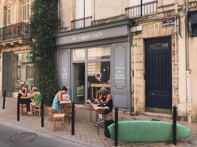 The grey painted 4e Vague Café storefront on Rue Notre Dame with outdoor tables, customers seated at a distance and a surfboard leaning against the wall