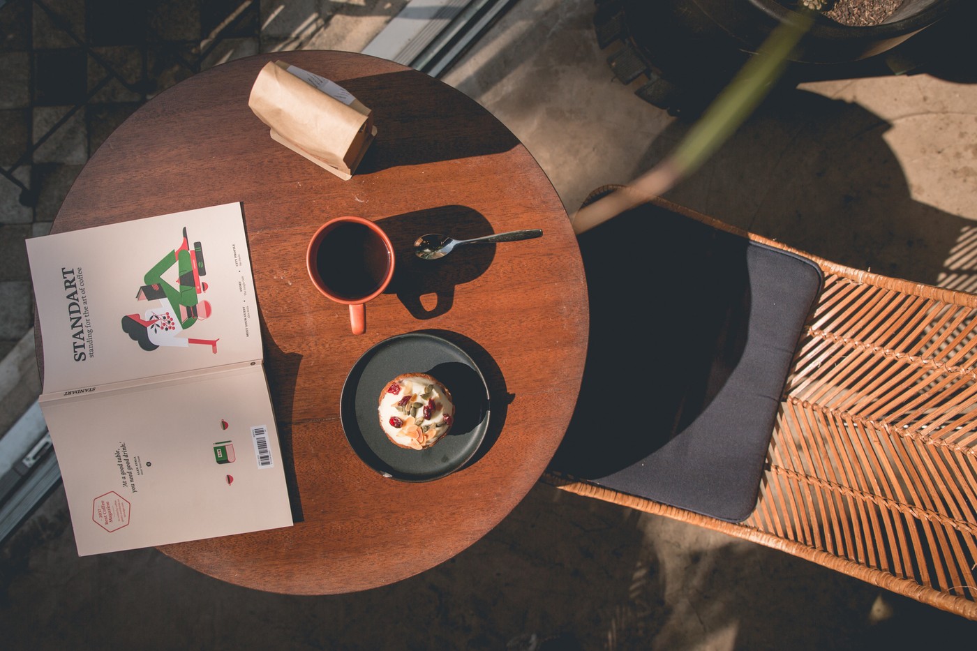 An overhead view of a terrace table with a red cup of black coffee, a small tart, a bag of beans and a coffee magazine
