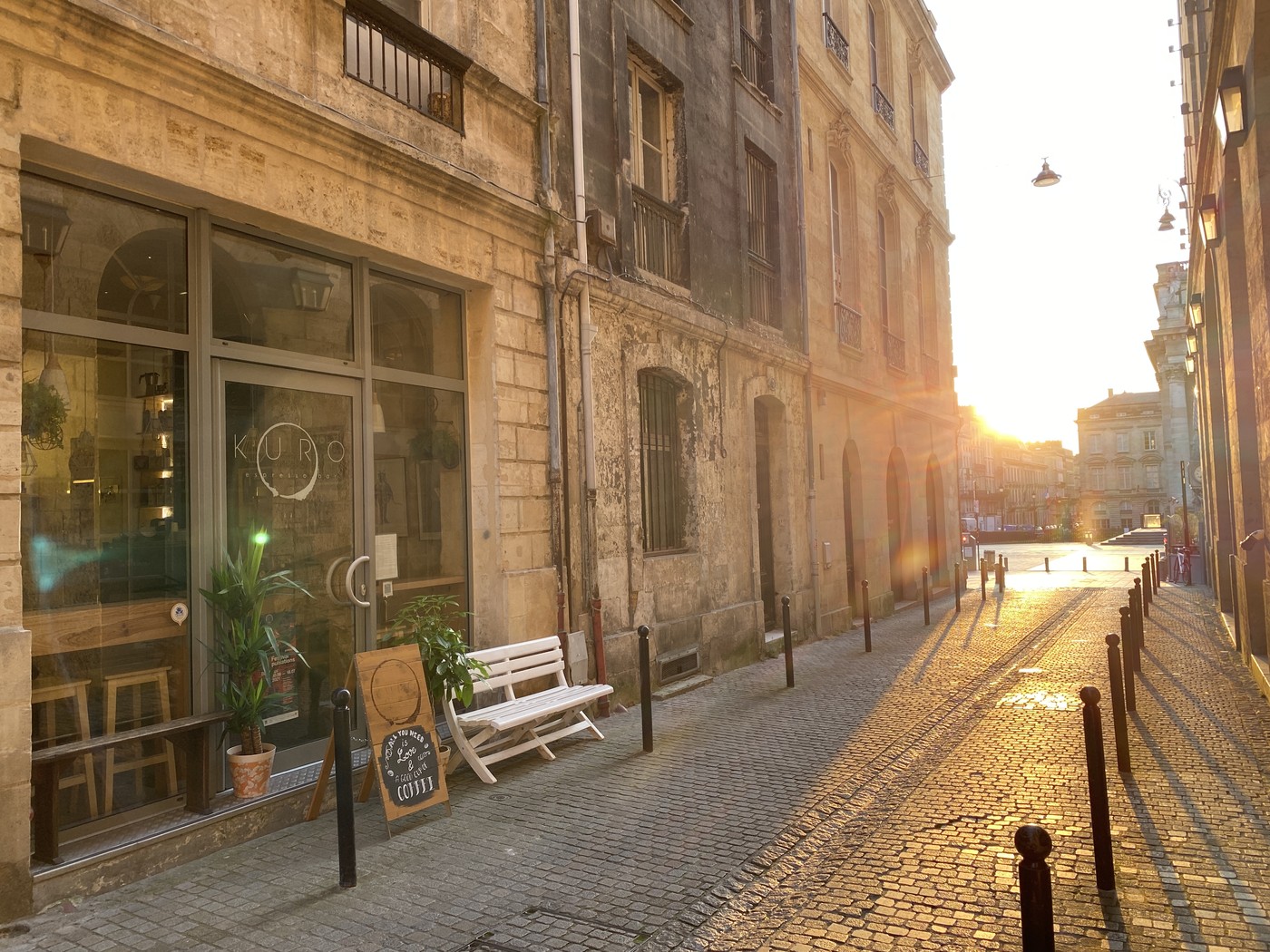 The KURO storefront on a cobblestoned lane at sunset, with a sandwich board reading All You Need Is Love And A Good Cup Of Coffee beside a white bench and potted plants