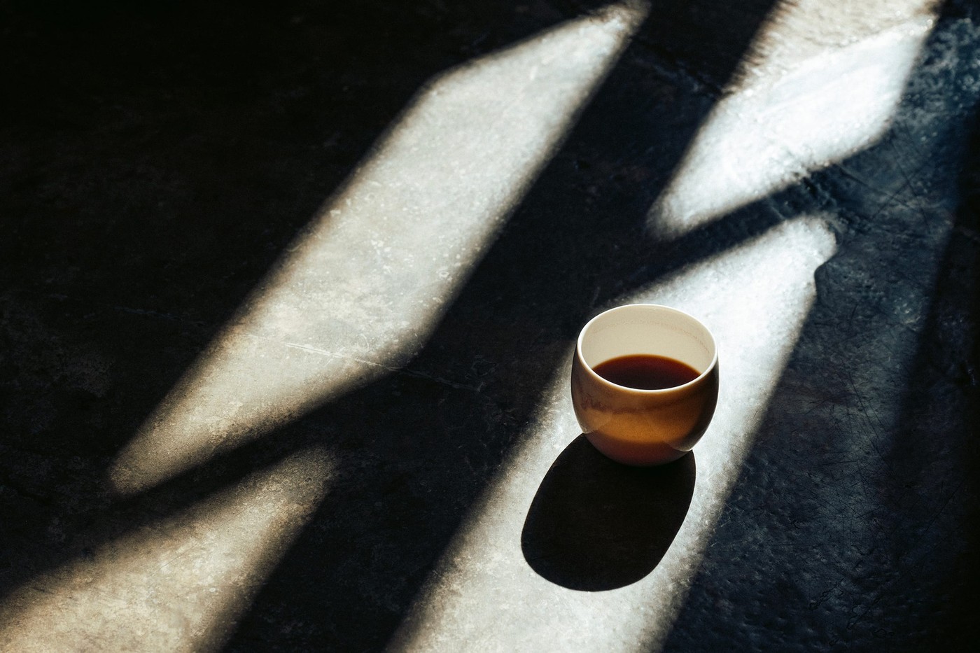 A small ceramic cup of black coffee sitting on a dark stone counter, lit by dramatic diagonal window light
