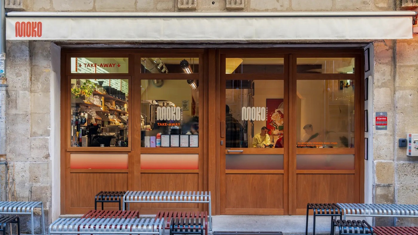 The Moko Coffee storefront at dusk, its retro orange logo and take away signage glowing behind wood framed windows with a bag of beans and shelving visible inside