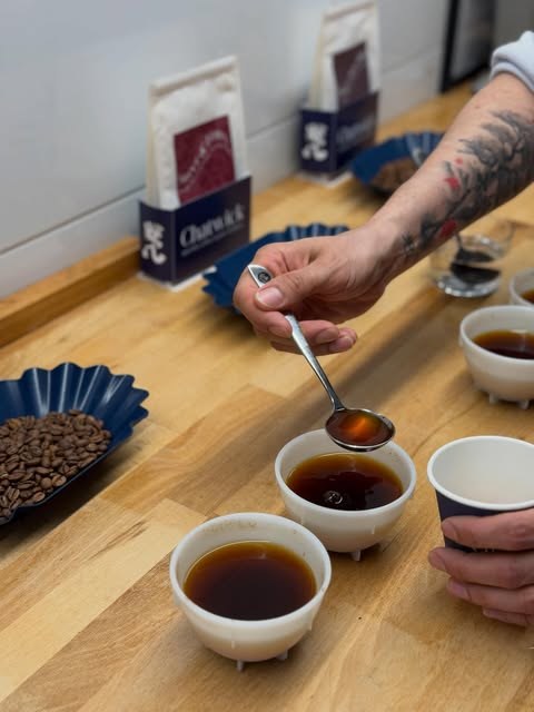A cupping session with Chatwick branded bags, roasted beans and cups of coffee on a wooden counter, a hand holding a cupping spoon
