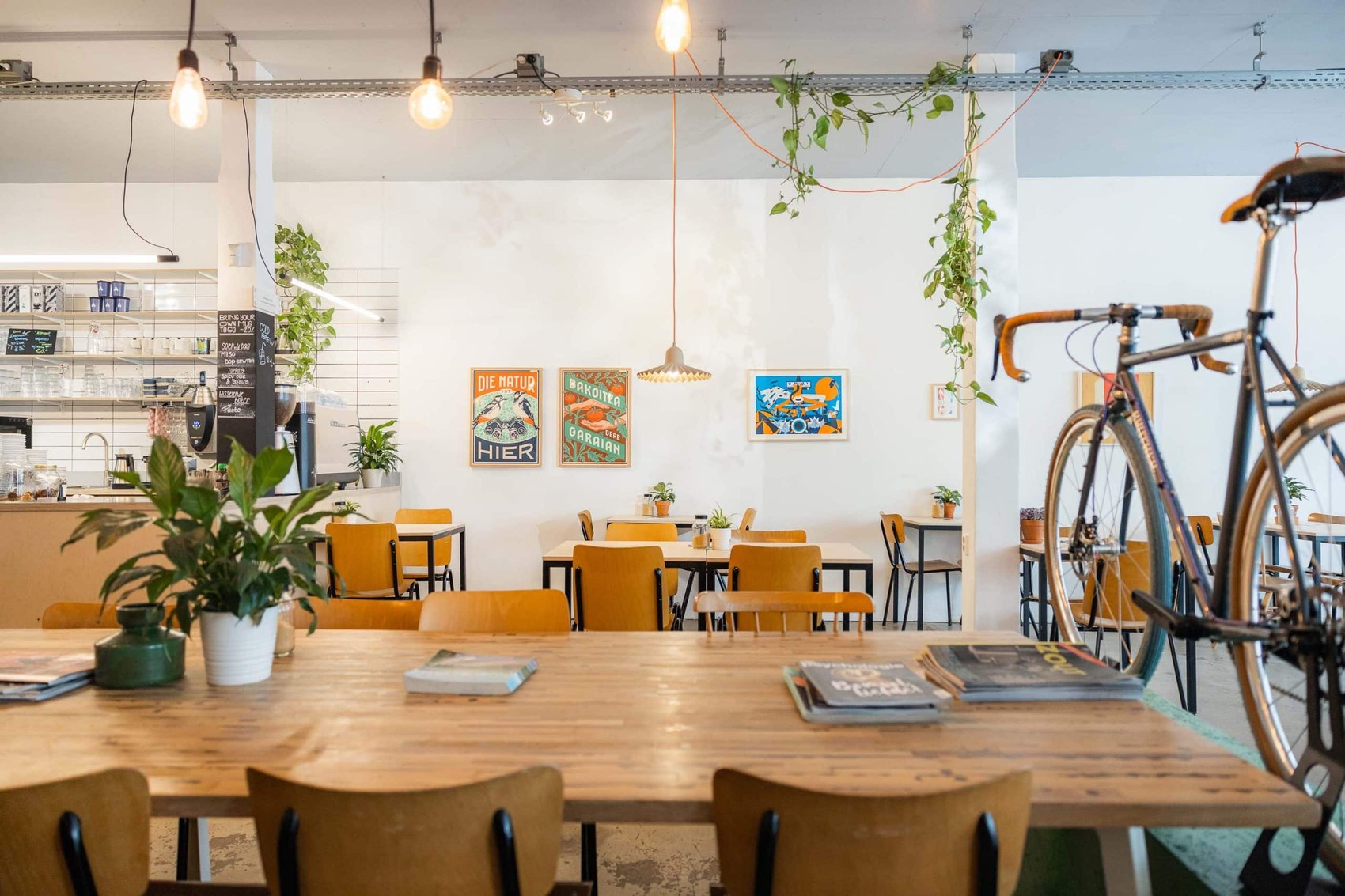 Wooden cafe tables and mismatched chairs beneath hanging plants and gallery posters, a road bike parked among the seating