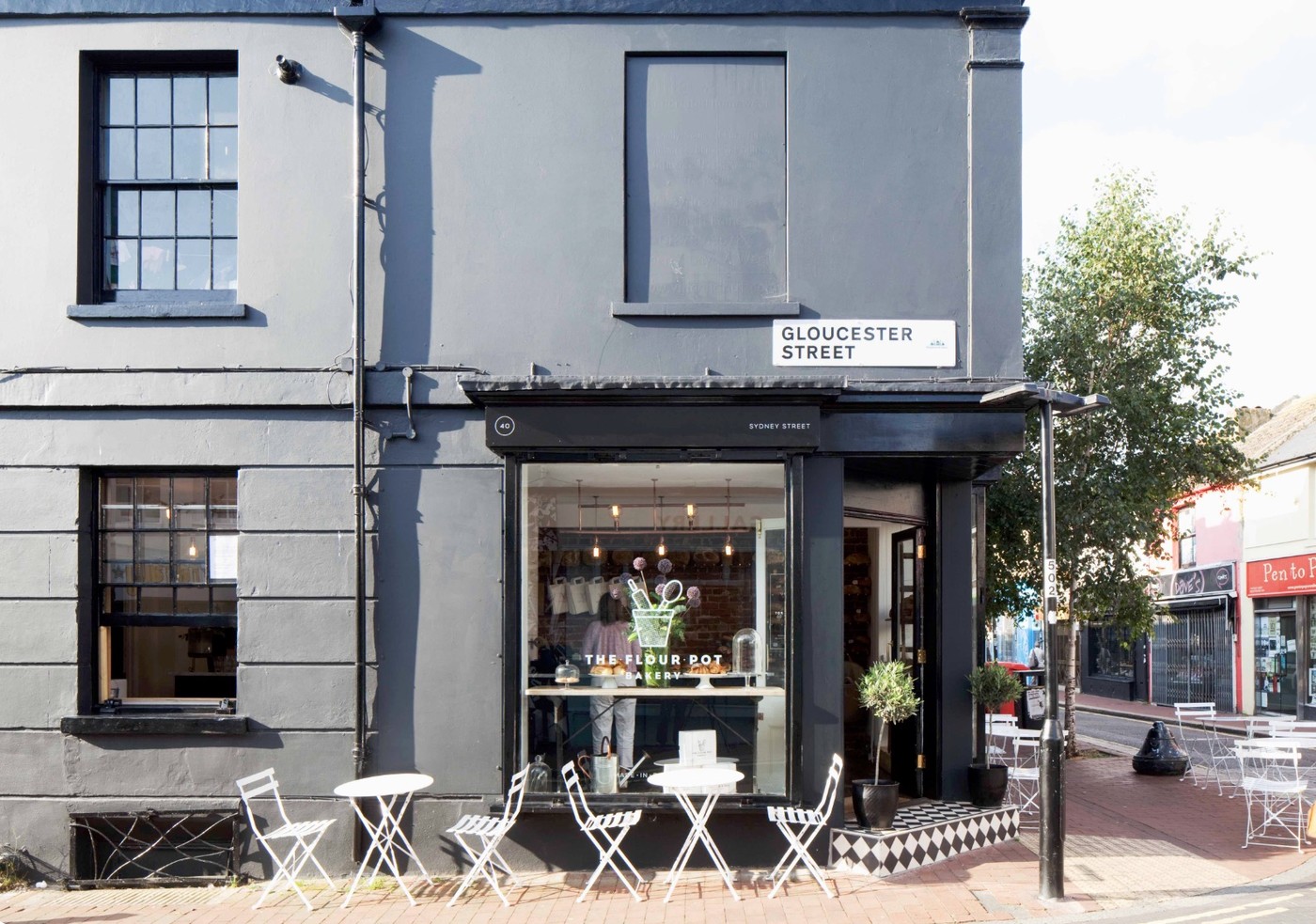 The black corner storefront of The Flour Pot Bakery on Sydney Street with white bistro tables out front