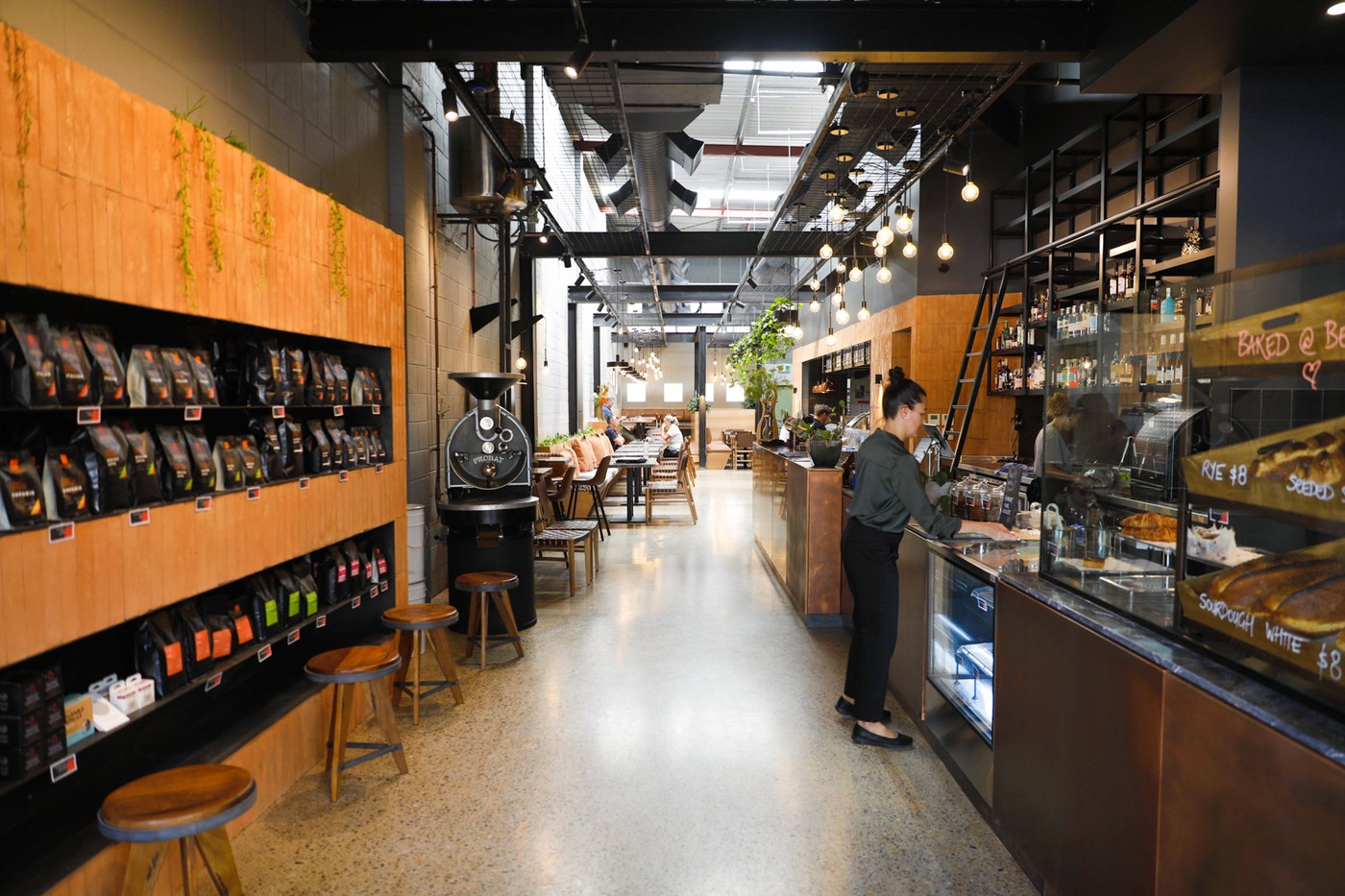 Interior of the Fortitude Valley roastery cafe, with bagged retail beans on wall shelving, a Probat roaster and a barista at the bar