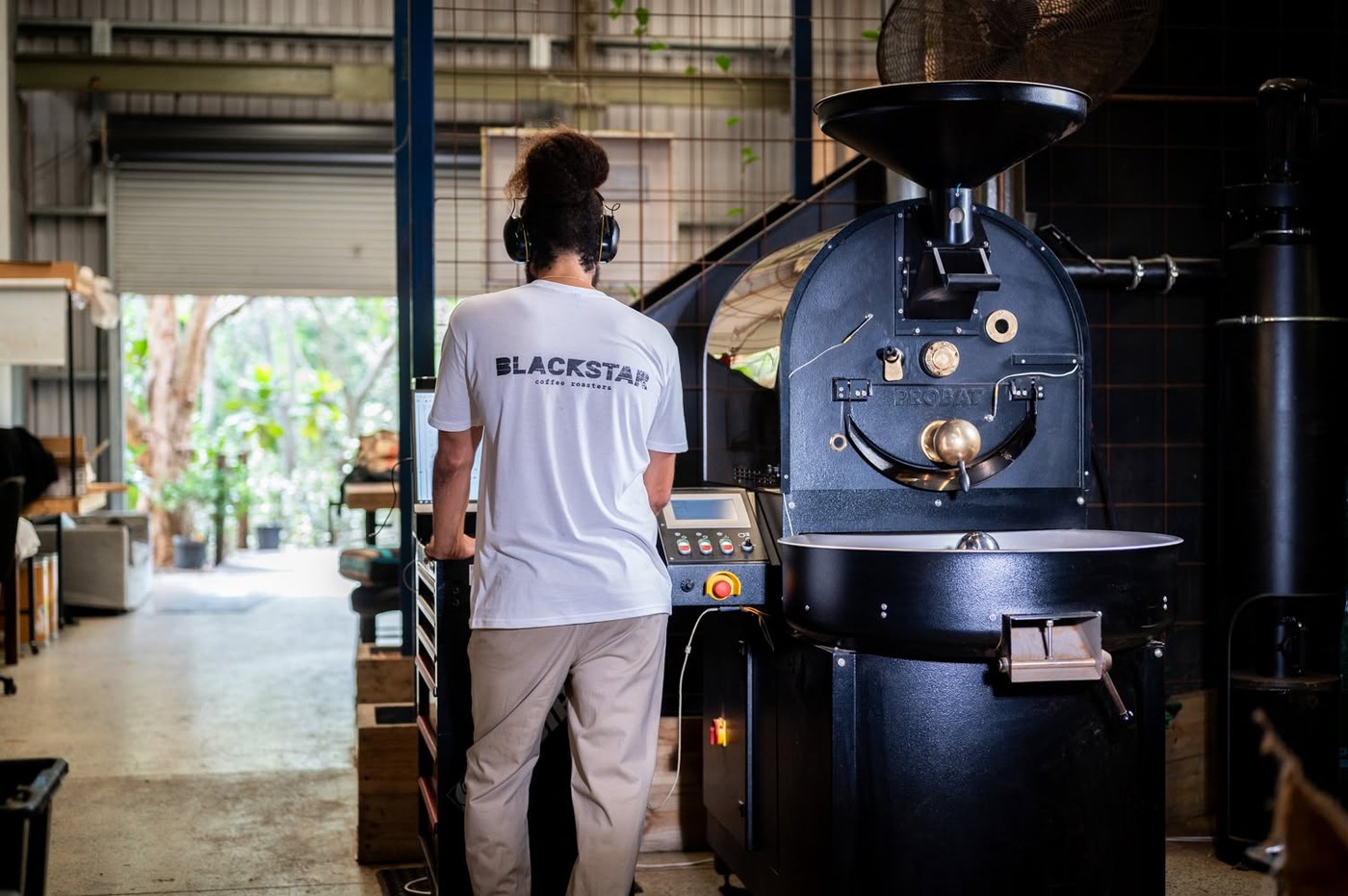 A roaster in a Blackstar branded shirt, back turned, standing beside the Probat roasting machine in the open sided warehouse