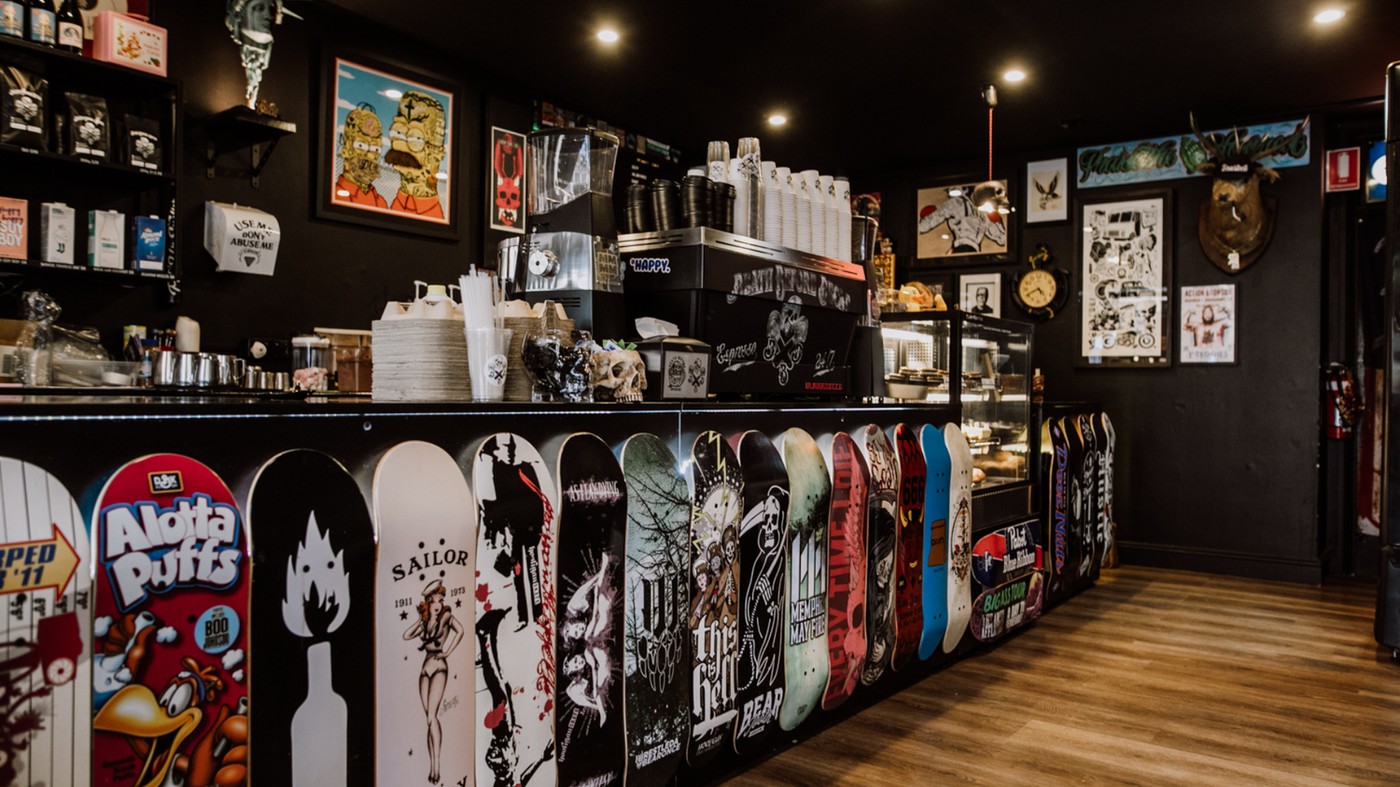 Skate decks lining the counter front below a Probat-branded espresso machine and band posters at Death Before Decaf