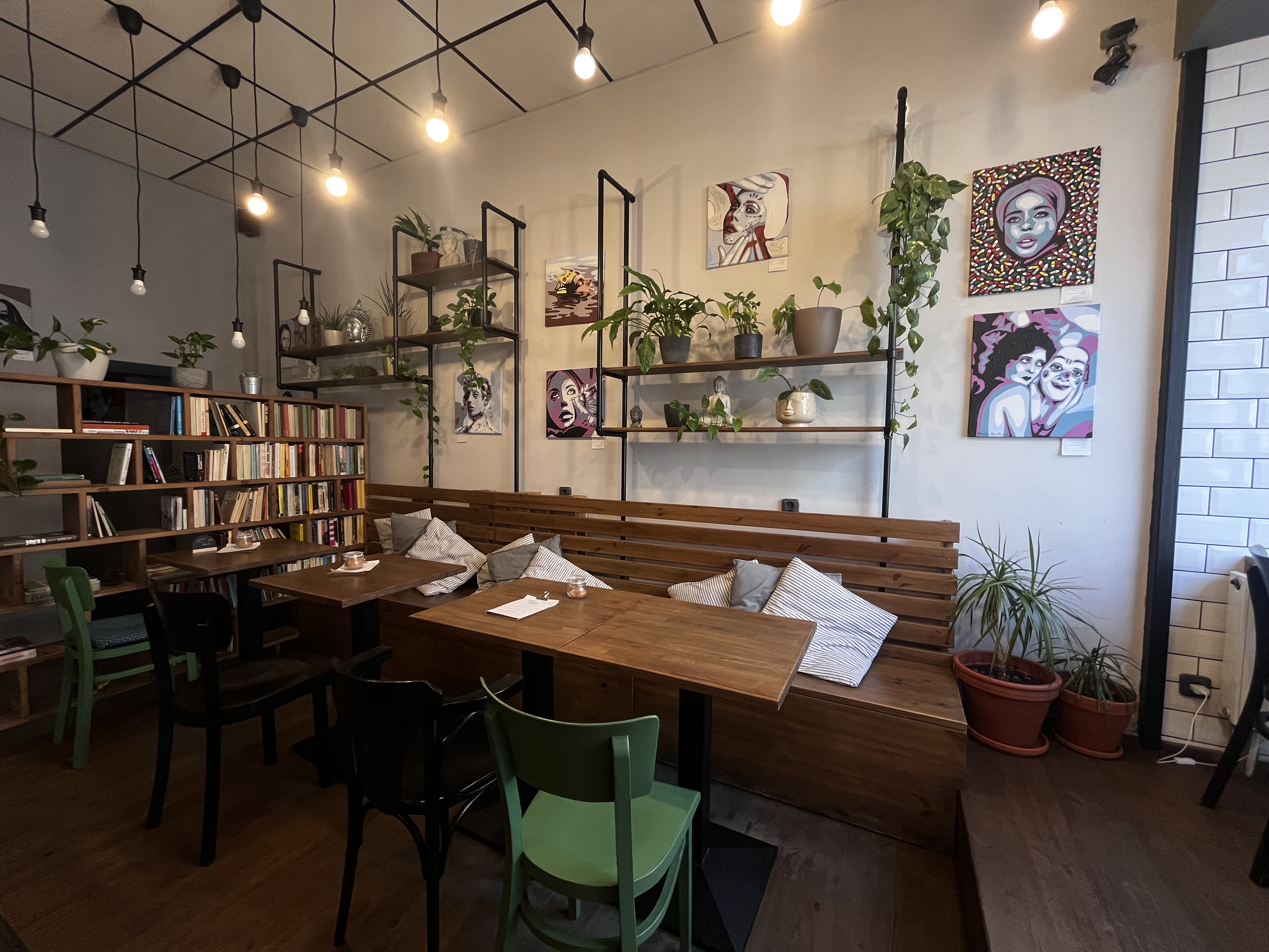 Coffee Bar Story's cozy interior with wooden bench seating and cushions, book-lined shelves, hanging plants, and pop-art portraits on a white wall under string lights