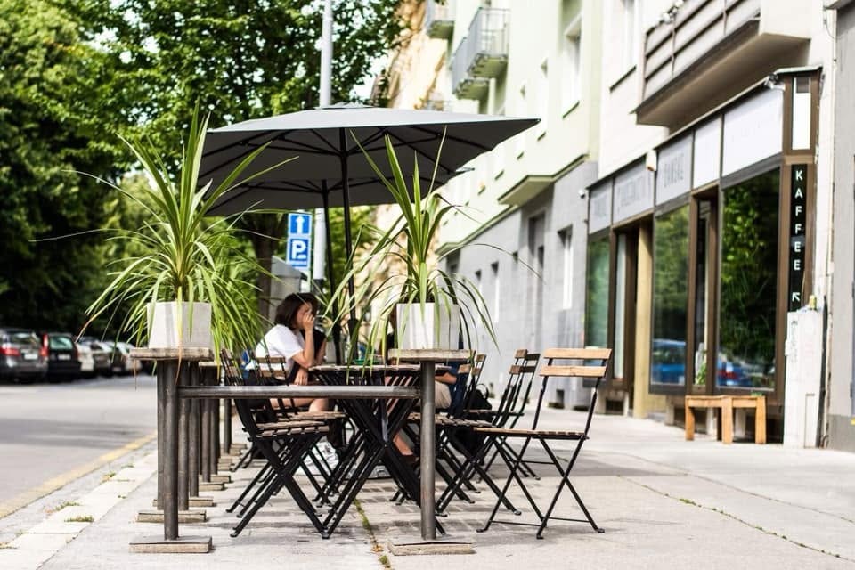 Outdoor seating under an umbrella and potted plants on the sidewalk outside Milady Kaffee